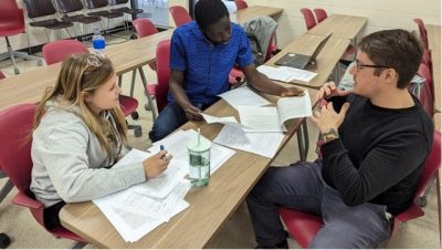 Julia Shapiro, Foster Agyei, and Chris Armstrong sit across from each other with papers strewn on the desk bfore them, talking about proposal ideas. Courtesy of Meryl Mims