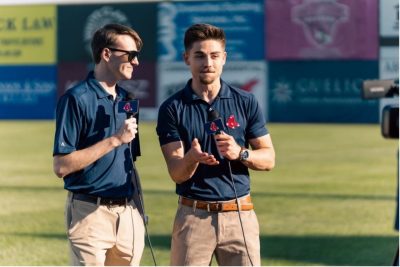 Two male students stand on a baseball field with microphones.