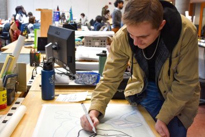 A student stands beside a table holding a marker that he is using to trace shapes on a paper lying on top of what looks to be a map on the table.